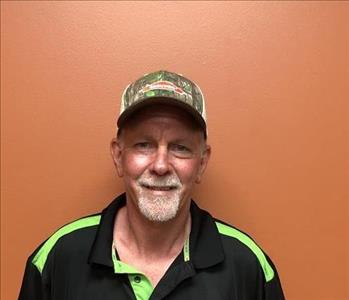 An older man in a black shirt with a SERVPRO logo stands against an orange background
