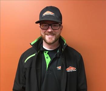 Man in a black hat and black and green SERVPRO Jacket stands against an orange background